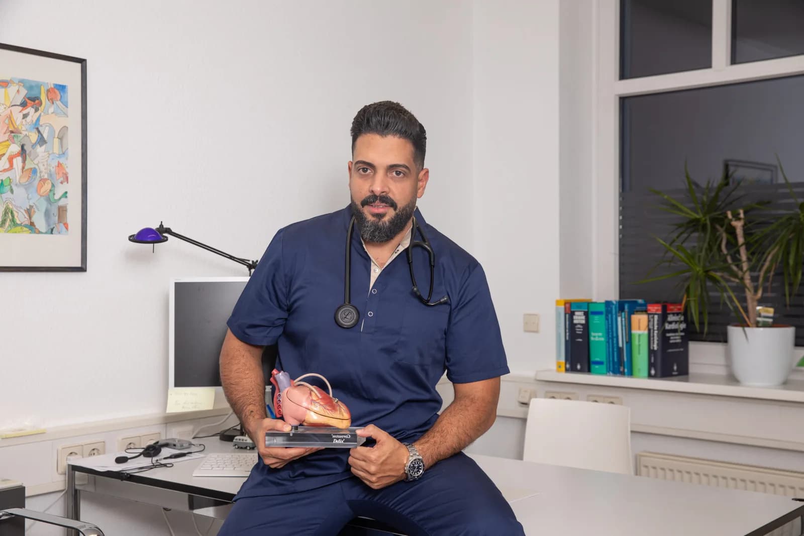 Dr. med. Karem Suleiman im Behandlungszimmer, mit Stethoskop und einem anatomischen Herzmodell in der Hand